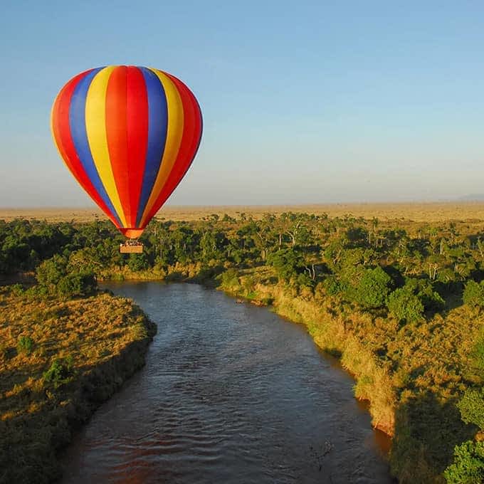 A hot-air balloon drifting over a river and the Maasai Mara plains at sunrise.