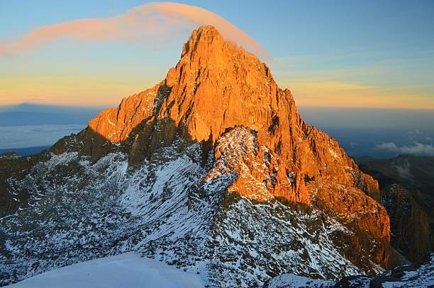 The rugged peak of Mount Kenya glowing orange at sunrise above snow and cloud.